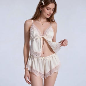 Könnte beinhalten: Ein cremefarbenes zweiteiliges Pyjama-Set bestehend aus einem Camisole und einer Shorts. Das Camisole verfügt über einen V-Ausschnitt mit zarter Spitzenborte, dünne Spaghettiträger und eine Bindeschleife vorne. Die Shorts hat einen Spitzenbesatz.