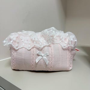 May include: A pink cosmetic bag with a white lace trim and a small white bow. The bag has a ribbed texture and a delicate, feminine design. The bag is designed to hold makeup and other beauty products.