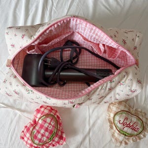 May include: A floral-patterned travel bag with a pink gingham interior, holding a dark-colored hair dryer. Two heart-shaped sachets, one with the name "Sophia" embroidered on it, are next to the bag. The bag and sachets are on a white surface.