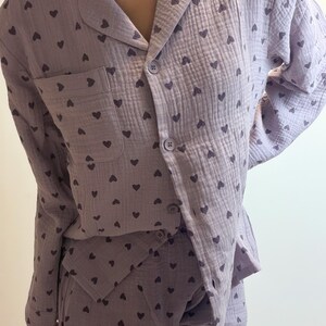 Op de afbeelding: Een lichtpaarse pyjamaset met een pyjamashirt met lange mouwen en een bijpassende broek, voorzien van een herhalend donkerpaars hartjespatroon. Het shirt heeft een reverskraag, een borstzakje en vier bijpassende knopen aan de voorzijde.