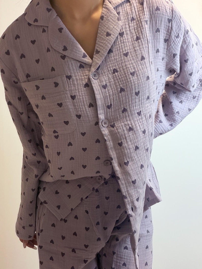 Op de afbeelding: Een lichtpaarse pyjamaset met een pyjamashirt met lange mouwen en een bijpassende broek, voorzien van een herhalend donkerpaars hartjespatroon. Het shirt heeft een reverskraag, een borstzakje en vier bijpassende knopen aan de voorzijde.