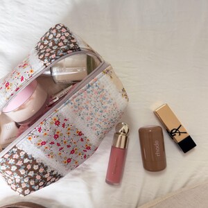 May include: A floral patterned cosmetic bag with a zipper, open to reveal various beauty products. A pink lip gloss, a brown tube, and a gold lipstick are next to the bag. The lipstick has a black accent.