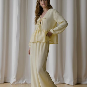 Könnte beinhalten: Ein hellgelbes zweiteiliges Pyjama-Set. Das Oberteil hat einen V-Ausschnitt mit Schleife, lange Ärmel und eine Schößchen-Taille. Die passende Hose ist weit geschnitten. Der Stoff hat ein strukturiertes, gekrepptes Aussehen.