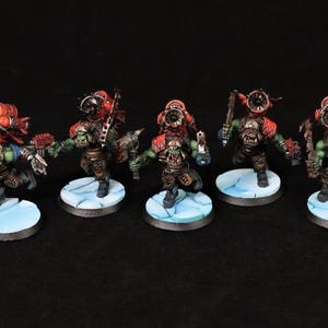 Puede incluir: Cinco figuras en miniatura de orcos de piel verde, cada uno con armadura roja, ropa marrón y armas. Se paran sobre bases circulares de color azul claro. Las figuras están pintadas con detalle, y el fondo es negro.