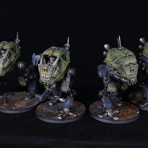 May include: Four miniature olive green and gray mechs with detailed weaponry and pilot figures. The mechs are posed on circular bases with a metallic texture. The image is set against a black background, highlighting the models.