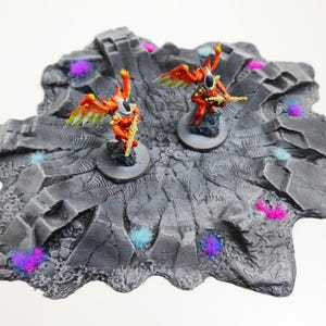 Könnte beinhalten: Zwei Miniaturfiguren mit orangefarbener Rüstung und gefiederten Flügeln stehen auf einer grauen, felsigen Geländebasis. Die Basis ist mit bunten, gesprenkelten Akzenten verziert. Die Figuren halten Waffen.