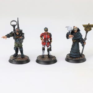 Puede incluir: Tres figuras en miniatura de personajes con trajes detallados. Una figura viste un uniforme verde y negro con una mochila de radio. Otra viste un uniforme rojo. La tercera figura viste una túnica oscura y sostiene un bastón, con un pájaro en el hombro.