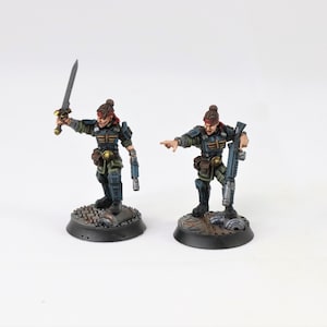 May include: Two miniature figurines of female soldiers, each with a unique pose. One holds a sword aloft, the other a rifle. Both are clad in dark green and blue armor, standing on detailed circular bases. The figures are painted with intricate detail.