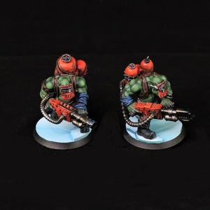 Könnte beinhalten: Zwei Miniaturfiguren von grün gehäuteten Kriegern, jeder bewaffnet mit einer roten Waffe und mit einem roten Helm und Rucksack. Sie stehen auf einer hellblauen Basis vor einem schwarzen Hintergrund. Die Figuren sind detailliert und bemalt.