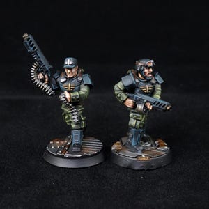 May include: Two miniature figurines of soldiers in olive green and blue uniforms, each holding a weapon. One figure carries a large machine gun with a belt of bullets, while the other holds a smaller rifle. The figures stand on circular bases.
