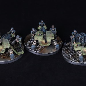 May include: Three miniature military vehicles with soldiers. The vehicles are painted in shades of green and brown, with detailed weaponry. The soldiers are wearing blue uniforms. The bases are circular with a metallic finish. The background is black.