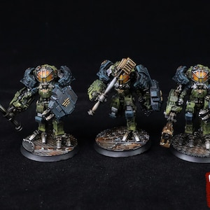 May include: Three miniature figurines of armored robots, each painted in shades of green, gray, and brown. They are equipped with weapons and shields, standing on circular bases. The robots have detailed features and a weathered appearance.