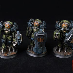 May include: Three miniature military robots with olive green and dark blue armor, orange visors, and weapons. One robot holds a shield. The robots stand on textured circular bases against a black background. The Creed's Painting logo is in the bottom right corner.