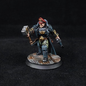 May include: Miniature figure of a military officer in a dark blue coat, red hat, and gold accents, holding a hammer and a pistol. The figure stands on a circular base with a textured surface.