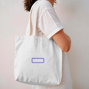 May include: A plain white canvas tote bag featuring a simple, thin-lined dark blue rectangular border printed in the center. The bag has two shoulder straps and is constructed from a lightweight, off-white fabric material with a clean design.