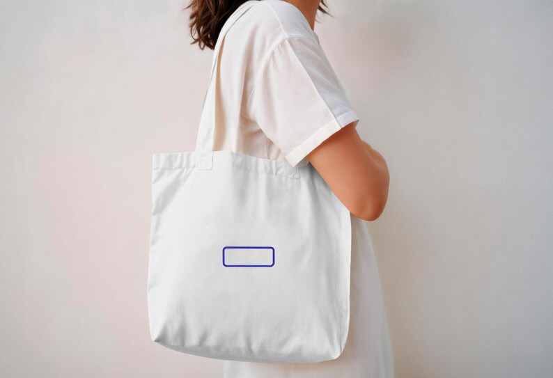 May include: A plain white canvas tote bag featuring a simple, thin-lined dark blue rectangular border printed in the center. The bag has two shoulder straps and is constructed from a lightweight, off-white fabric material with a clean design.