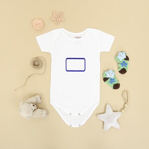 May include: A white short-sleeved baby bodysuit featuring a blank rectangular blue frame on the chest. The bodysuit is surrounded by a small beige teddy bear, a star-shaped rattle, a coil of twine, and two green socks with star patterns.