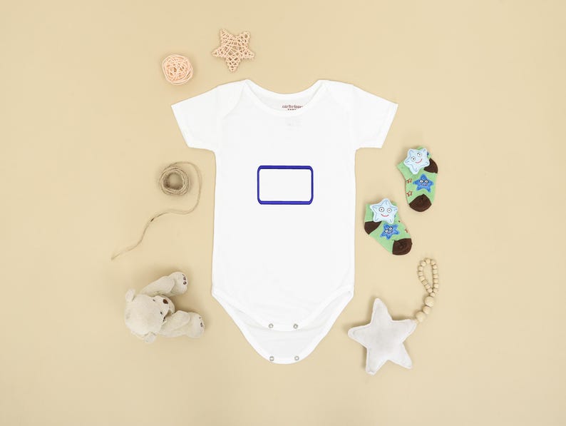 May include: A white short-sleeved baby bodysuit featuring a blank rectangular blue frame on the chest. The bodysuit is surrounded by a small beige teddy bear, a star-shaped rattle, a coil of twine, and two green socks with star patterns.