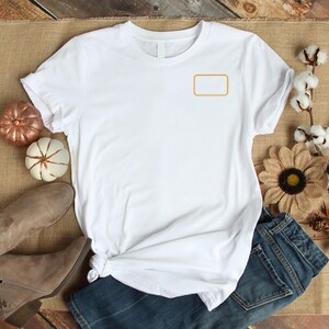 May include: A white short-sleeved t-shirt featuring a blank, rectangular orange outline on the upper left chest area. The shirt has a crew neckline and is displayed flat with a knotted hem detail on the lower left side of the garment.