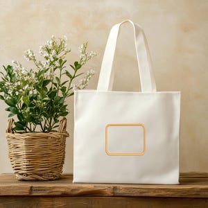 May include: A white canvas tote bag featuring a rectangular patch with an orange border centered on the front. The bag has two sturdy shoulder straps and a clean, minimalist design, providing a blank space for customization or branding.