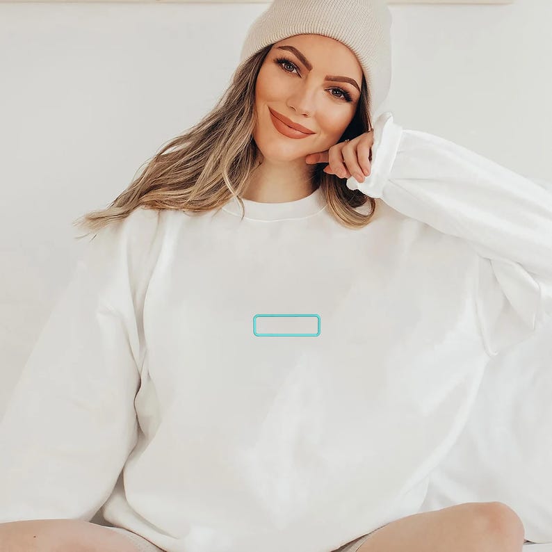 May include: A solid white long-sleeve crewneck sweatshirt featuring a small, rectangular teal outline graphic centered on the chest. The garment is paired with a matching light beige knit beanie cap, creating a clean and simple monochromatic look.