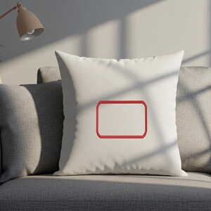 May include: A square off-white throw pillow featuring a centered, empty rectangular frame embroidered with a bold red thread outline. The pillow has a smooth, solid-colored fabric surface and is positioned upright, showcasing the stitched design.
