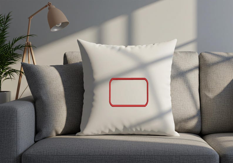 May include: A square off-white throw pillow featuring a centered, empty rectangular frame embroidered with a bold red thread outline. The pillow has a smooth, solid-colored fabric surface and is positioned upright, showcasing the stitched design.