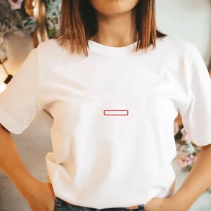 May include: A plain white short-sleeved t-shirt featuring a small, thin, rectangular red outline graphic centered on the chest. The garment has a classic crew neckline and a standard fit, constructed from a smooth, opaque white fabric.