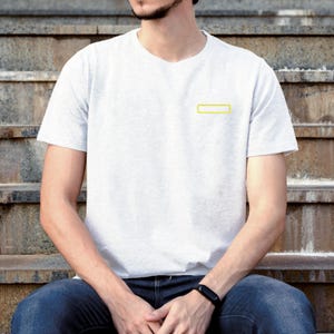 May include: A light gray short-sleeved t-shirt featuring a small, rectangular yellow outline graphic on the left chest. The shirt has a crew neckline and a plain, solid-colored back, providing a simple and clean appearance for casual wear.