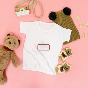 May include: A plain white short-sleeved t-shirt featuring a red rectangular outline on the chest, accompanied by a brown teddy bear, a mustard yellow knit beanie with two black pom-poms, a toy camera, and two small socks with bear faces.