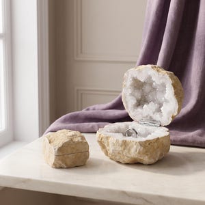 May include: A geode ring box with a rough, tan exterior and a hollow, crystalline interior featuring white, jagged quartz-like formations. The box is hinged with a silver-toned metal mechanism and contains a simple, thin silver-colored metal ring.