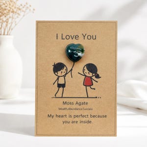 May include: A rectangular kraft paper card featuring the text I Love You at the top and a cartoon illustration of two figures holding a string attached to a heart-shaped dark green moss agate stone. Below, the text reads Moss Agate, Wealth.Abundance.Success, and My heart is perfect because you are inside.