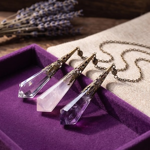 May include: Three pointed crystal pendants with ornate bronze-toned metal caps. The crystals are clear, pale pink, and purple. Each pendant is attached to a thin bronze-toned metal chain, resting together in a deep purple velvet-lined tray.