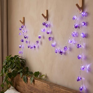 Könnte beinhalten: Eine LED-Lichterkette mit durchscheinenden, violetten, steinartigen Abdeckungen. Die Lichter sind über drei hölzerne Wandhaken drapiert und strahlen aus jedem einzelnen steinartigen Gehäuse entlang des Drahtes ein sanftes violettes Licht aus.