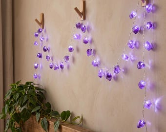 May include: A string of LED fairy lights featuring translucent purple, rock-shaped covers. The lights are draped across three wooden wall hooks, emitting a soft purple glow from within each individual stone-like housing along the wire.