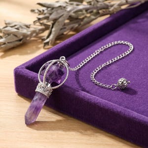 May include: A purple amethyst crystal pendulum featuring a faceted point and a star-shaped top encased in a silver-toned metal cage. The pendulum is attached to a silver-toned chain that ends with a small, textured spherical metal bead.
