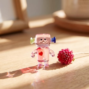 May include: A small, clear glass robot figurine with a pink-tinted body, a red heart detail on its chest, and small light green and dark blue accents on the sides of its head. The robot has a simple face with two black eyes and a curved mouth.