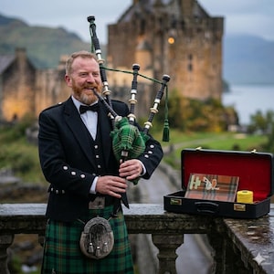 Puede incluir: Un hombre con vestimenta escocesa tradicional toca la gaita. Viste una chaqueta negra, chaleco y pajarita, con una falda escocesa a cuadros verdes y negros. Una maleta negra con tela escocesa y una bobina de hilo amarillo está cerca, con un castillo al fondo.
