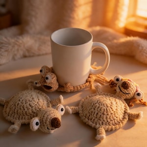 Puede incluir: Una taza de cerámica blanca descansa sobre un posavasos de ganchillo beige con forma de perezoso de dibujos animados. Se ven dos posavasos de perezoso adicionales. Los posavasos tienen ojos grandes y narices marrones.