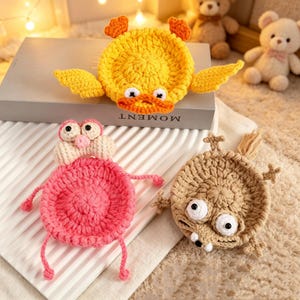May include: Three handmade crochet coasters in the shape of cartoon animals. A yellow duck with orange accents, a pink pig with a cream-colored head, and a tan creature with large eyes. The word "MOMENT" is visible on a book in the background.