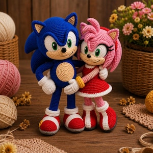 Może przedstawiać: Dwie szydełkowe lalki amigurumi przedstawiające Sonica i Amy Rose. Sonic jest niebieski z beżowym pyszczkiem oraz czerwono-białymi butami sportowymi. Amy Rose jest różowa, ma czerwoną sukienkę z białym wykończeniem oraz czerwono-białe buty.