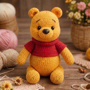 May include: A yellow crocheted bear toy with a round head, small ears, and black eyes. The bear is wearing a short-sleeved red shirt that covers its upper body. The texture of the yarn is visible throughout the entire surface of the stuffed animal.