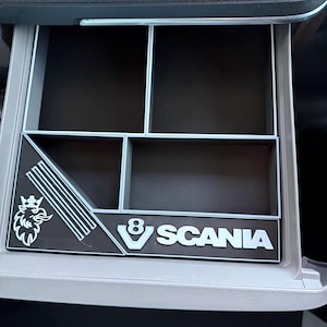 Op de afbeelding: Een zwarte kunststof opbergbak voor het dashboard met meerdere vakken, voorzien van een wit Scania-logo, een V8-embleem en een gestileerde griffioenafbeelding op de voorrand. De organizer is ontworpen voor een voertuigconsole met een lichtgrijs frame.