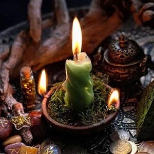 May include: A green candle, alight, is set in a small bowl of moss, with two other candles burning nearby. The scene includes a small pyramid, coins, and rune stones, creating a mystical aesthetic. A decorative container and small bottles are also present.
