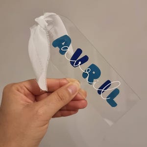 May include: A clear acrylic bookmark with the word "AVRIL" in blue and white script. The bookmark has a white ribbon attached at the top. The word "AVRIL" is written in a playful font.