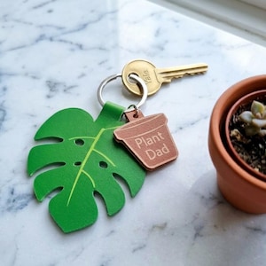 Op de afbeelding: Een groene leren monstera blad sleutelhanger met een bruin plantenpot tag met de tekst "Plant Dad". Een gouden sleutel is bevestigd aan de sleutelring. Een kleine terracotta pot met een vetplant staat op de achtergrond.