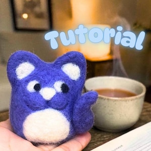 May include: A handmade, blue felted cat figurine with white accents, held in a hand. The cat has a white belly and ears, with black eyes. The word "Tutorial" is written above the cat in a light blue font.