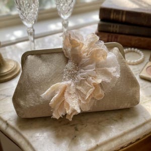 Op de afbeelding: Goudkleurige clutch met een getextureerd oppervlak. Een grote, crèmekleurige stoffen bloem met parelaccenten siert de voorkant. De rechthoekige clutch met sluiting is geschikt voor formele evenementen.