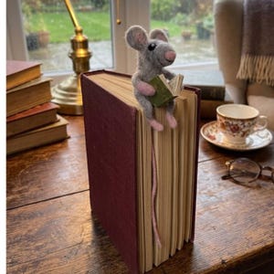 May include: A handmade felt mouse figurine reading a small green book, perched on the edge of a large, burgundy-covered book. The mouse has gray fur, pink ears and feet. A whimsical bookmark for book lovers.
