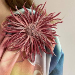 May include: A large, circular, dusty rose-colored felted flower brooch with long, thin, radiating petals and a textured, beaded center. The flower is attached to a soft, draped fabric featuring a gradient of pastel orange, pink, and blue tones.